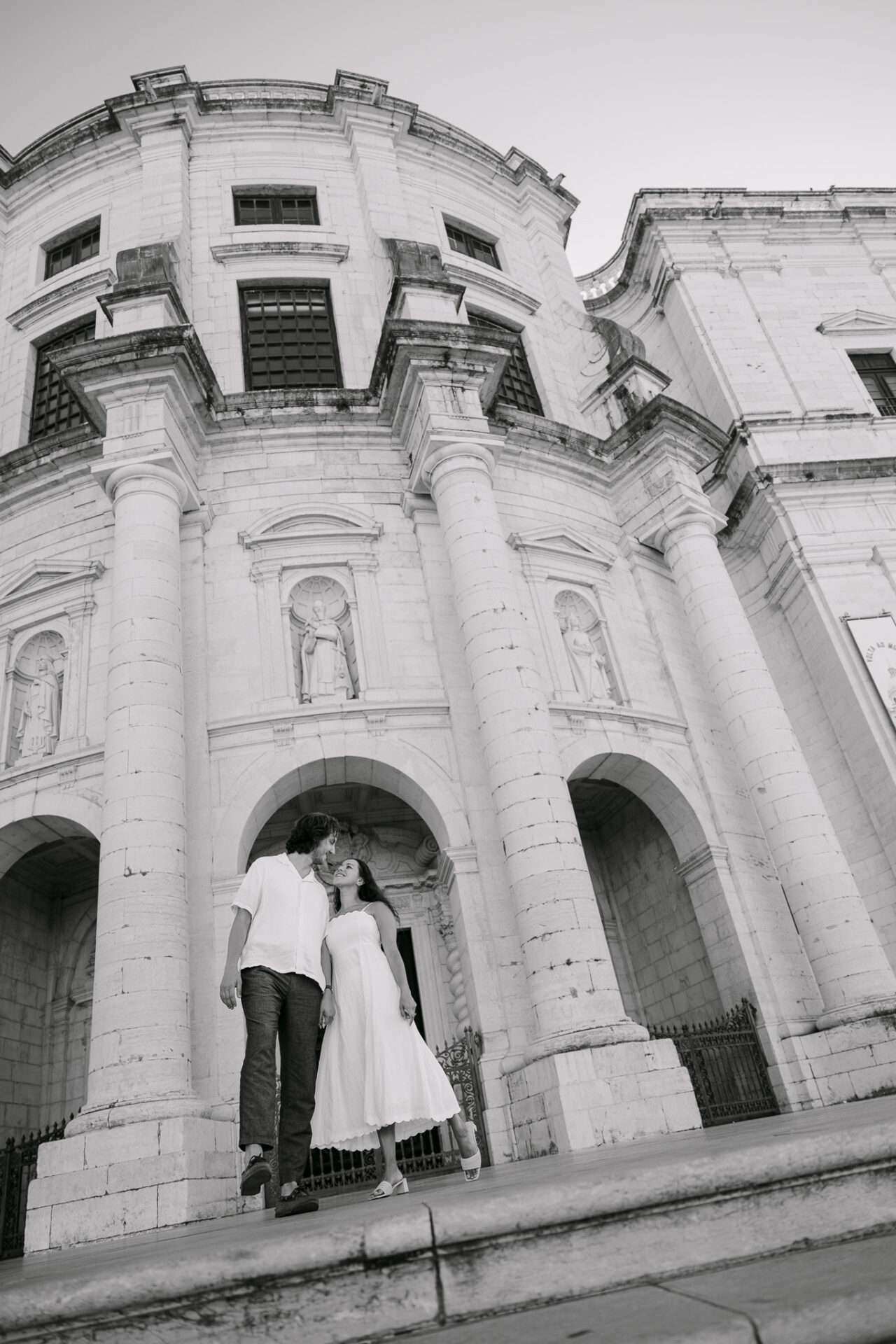 A couple stands close together, looking at each other, in front of a large, historic stone building with columns and statues in Lisbon—a timeless black and white moment, possibly capturing a heartfelt proposal from a low angle.