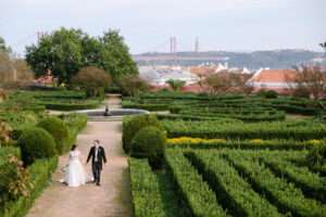 A bride and groom walk hand-in-hand along a garden path at Vandelli Botanical Garden, surrounded by manicured hedges, with a cityscape and the iconic red bridge of Lisbon wedding photography in the background.