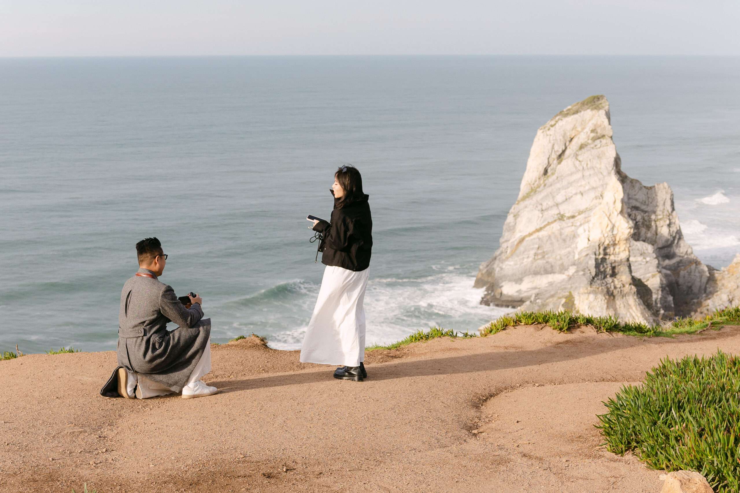 A man kneels on a cliff path holding a camera, facing a woman with another camera. Behind them, the ocean and a large rocky formation stretch under a clear sky—an idyllic scene for any engagement photographer in Portugal.
