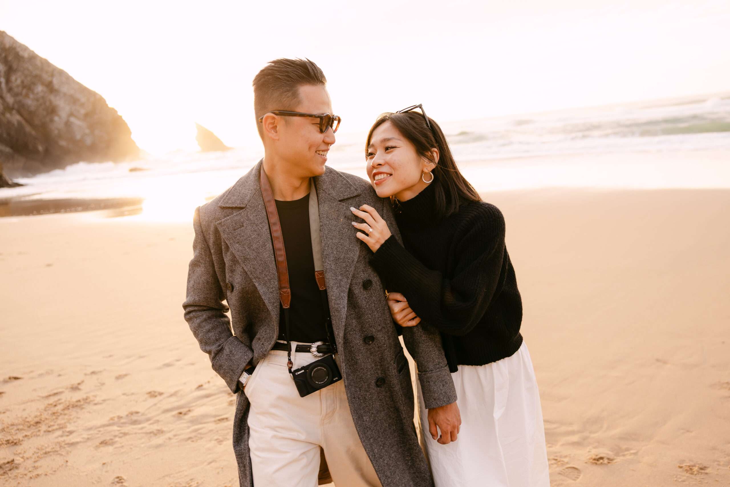 A smiling couple walks on a sandy beach at sunset, captured by an engagement photographer in Portugal. He wears sunglasses, a gray coat, and a camera; she, in a black sweater and white skirt, holds his arm as waves and cliffs complete the romantic scene.