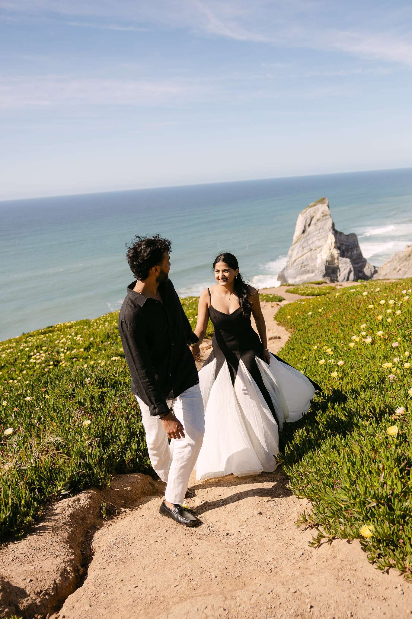 A couple walks along a coastal path surrounded by greenery, captured beautifully by an engagement photographer in Portugal. The woman wears a black and white dress, the man, a black shirt and white pants. Ocean waves and a large rock formation frame the scene.