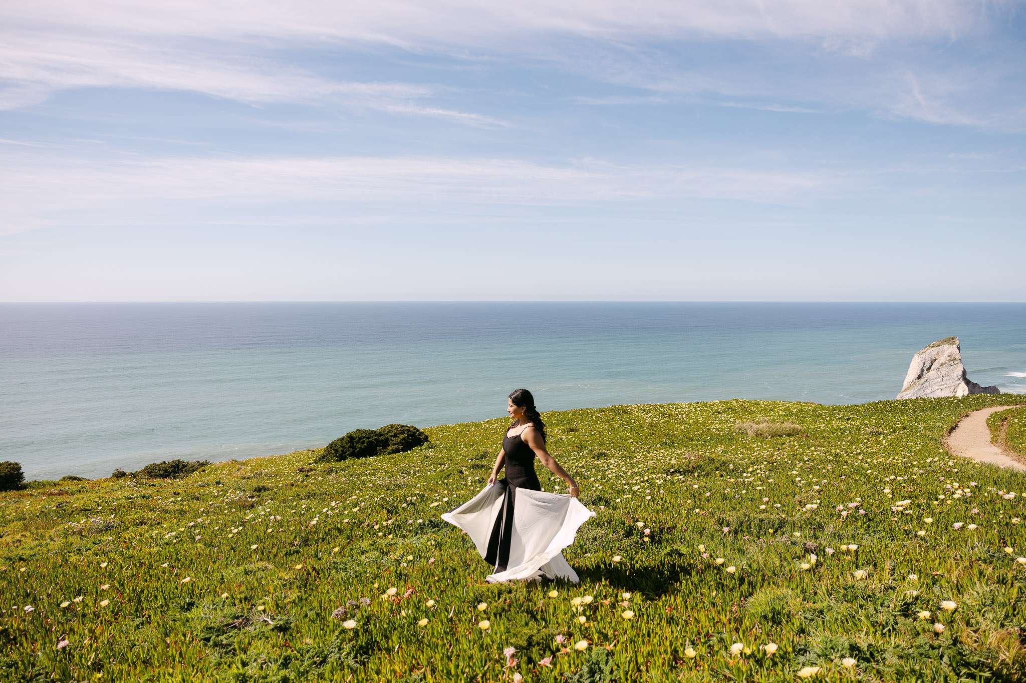 A woman in a flowing dress stands on a grassy field with wildflowers, overlooking the ocean under a blue sky. Perfect scenery for an engagement photographer in Portugal, with rocky cliffs and stunning coastline in the background.