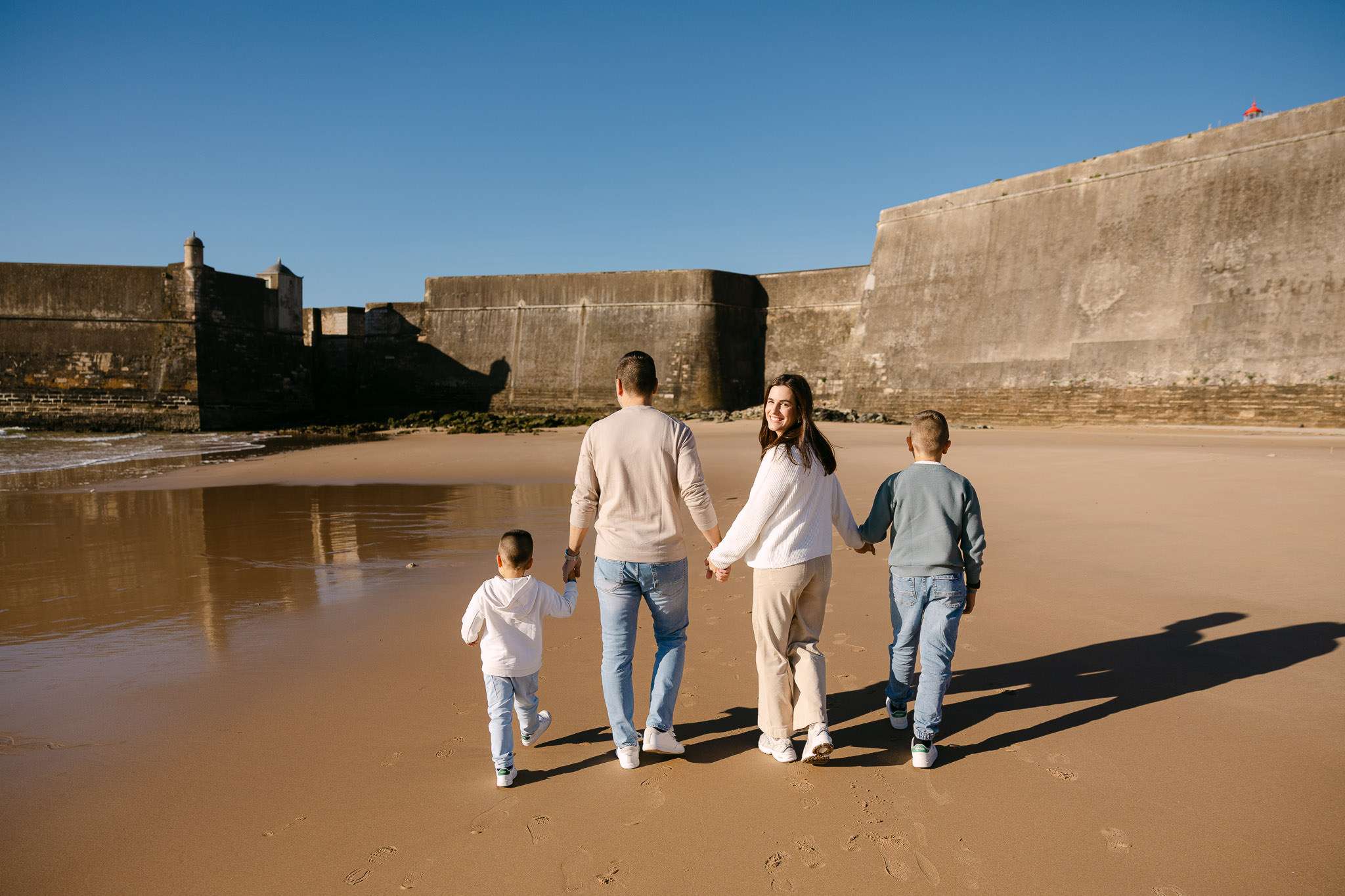 A family of four walks hand-in-hand along a sandy beach near large stone fort walls under a clear blue sky. The parents and two children are seen from behind, enjoying a sunny day together.