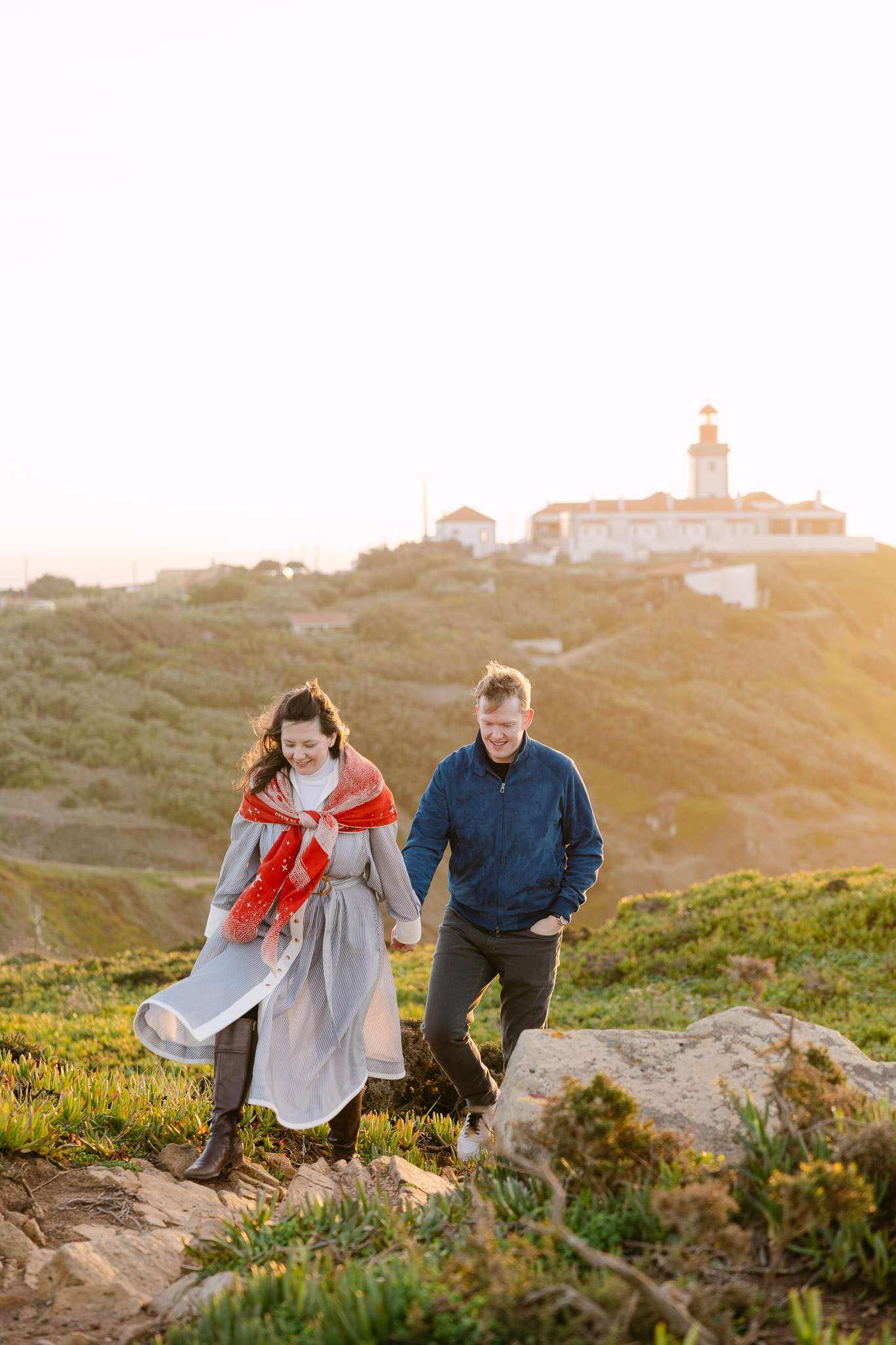 A couple walks hand in hand on a grassy hill at sunset, with a lighthouse and buildings behind them—captured beautifully by an engagement photographer in Portugal. The woman wears a gray dress and red scarf; the man is in a blue jacket and dark pants.