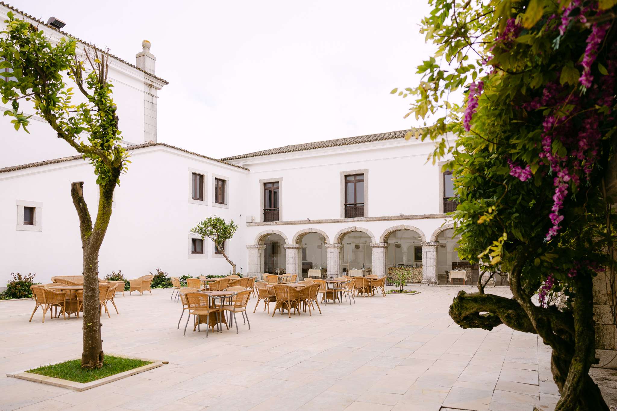 A spacious courtyard at Pousada do Castelo de Palmela, with white stone buildings, arched doorways, and large windows. Scattered tables and chairs amid green trees and purple flowers create a peaceful wedding setting.