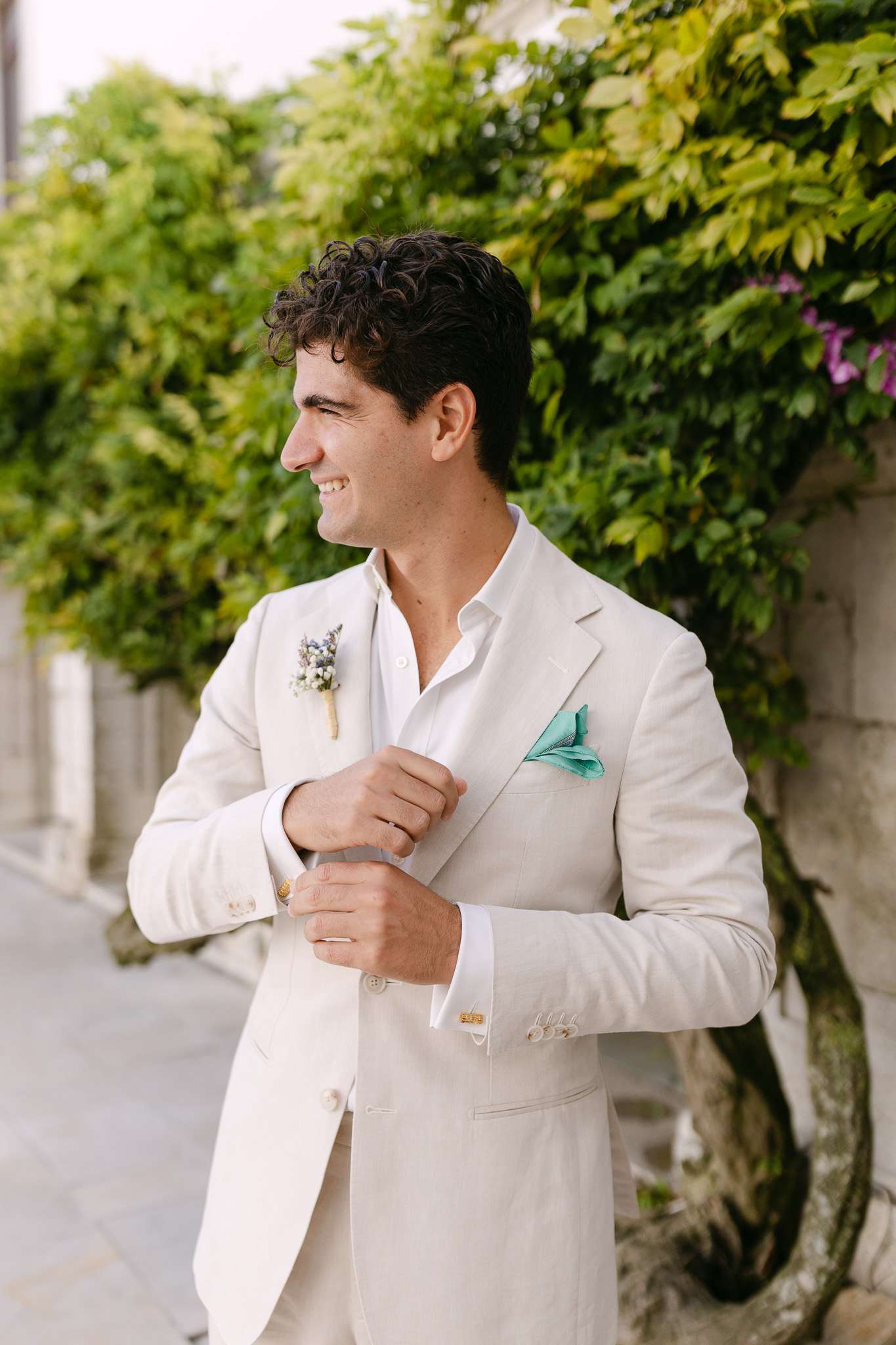 A man in a light beige suit adjusts his teal pocket square while smiling and looking to the side, standing outdoors near a wall covered with lush green foliage at a Pousada do Castelo de Palmela wedding.