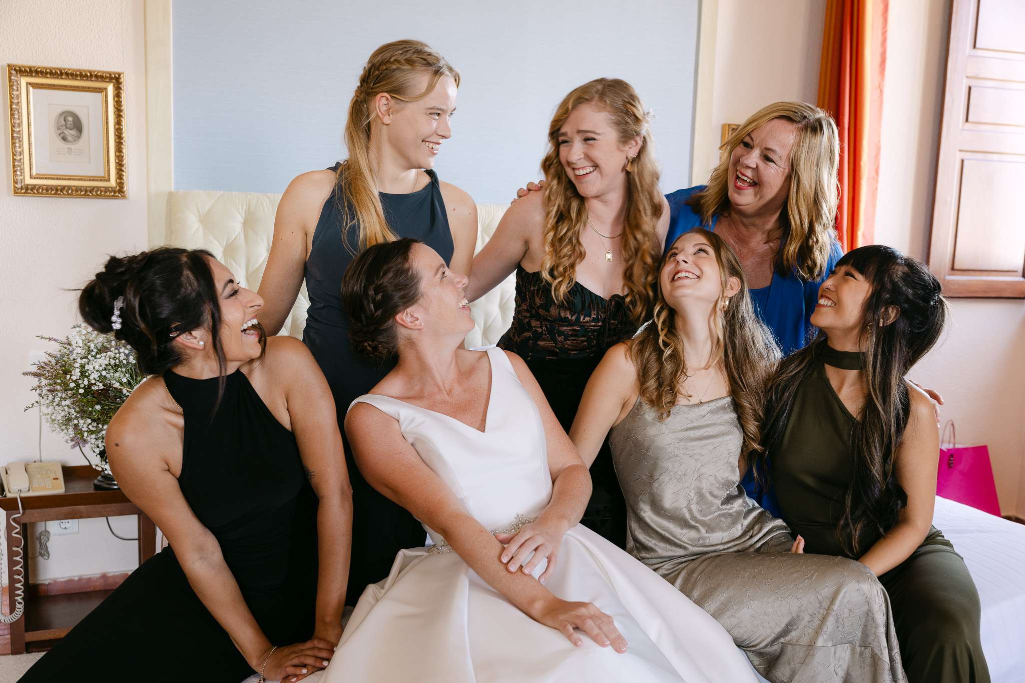 Six women in formal dresses sit and stand together on a bed, smiling and laughing in a brightly lit room at a pousada do castelo de palmela wedding, with framed photos on the wall and a wooden window in the background.