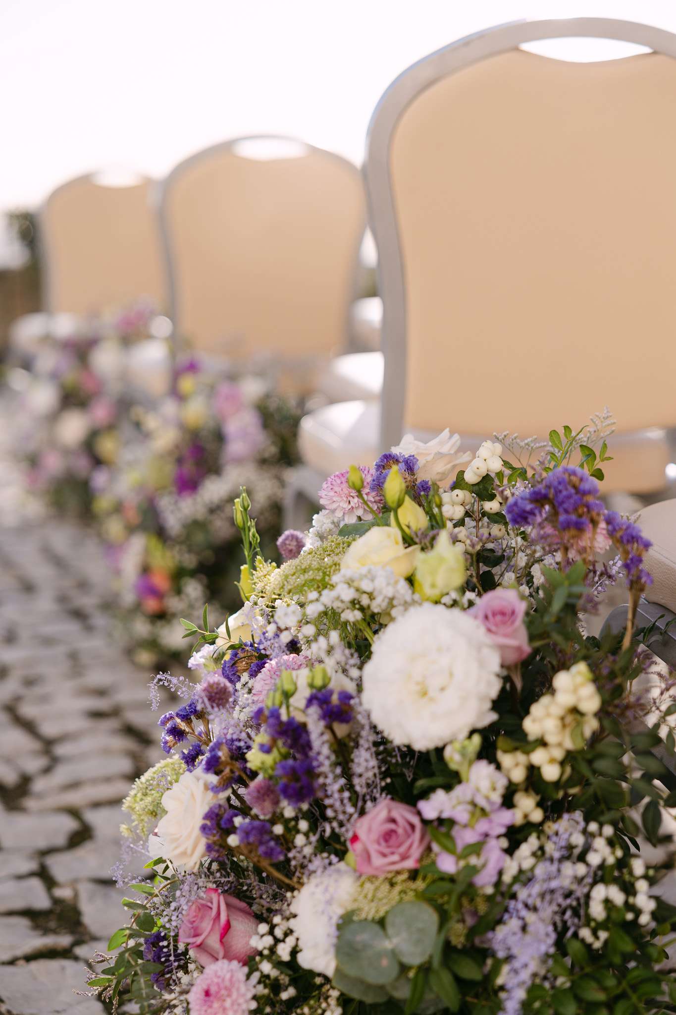 Rows of light-colored chairs are lined up outdoors on a stone path at a Pousada do Castelo de Palmela wedding, each decorated with lush floral arrangements featuring purple, white, and pink flowers with greenery.