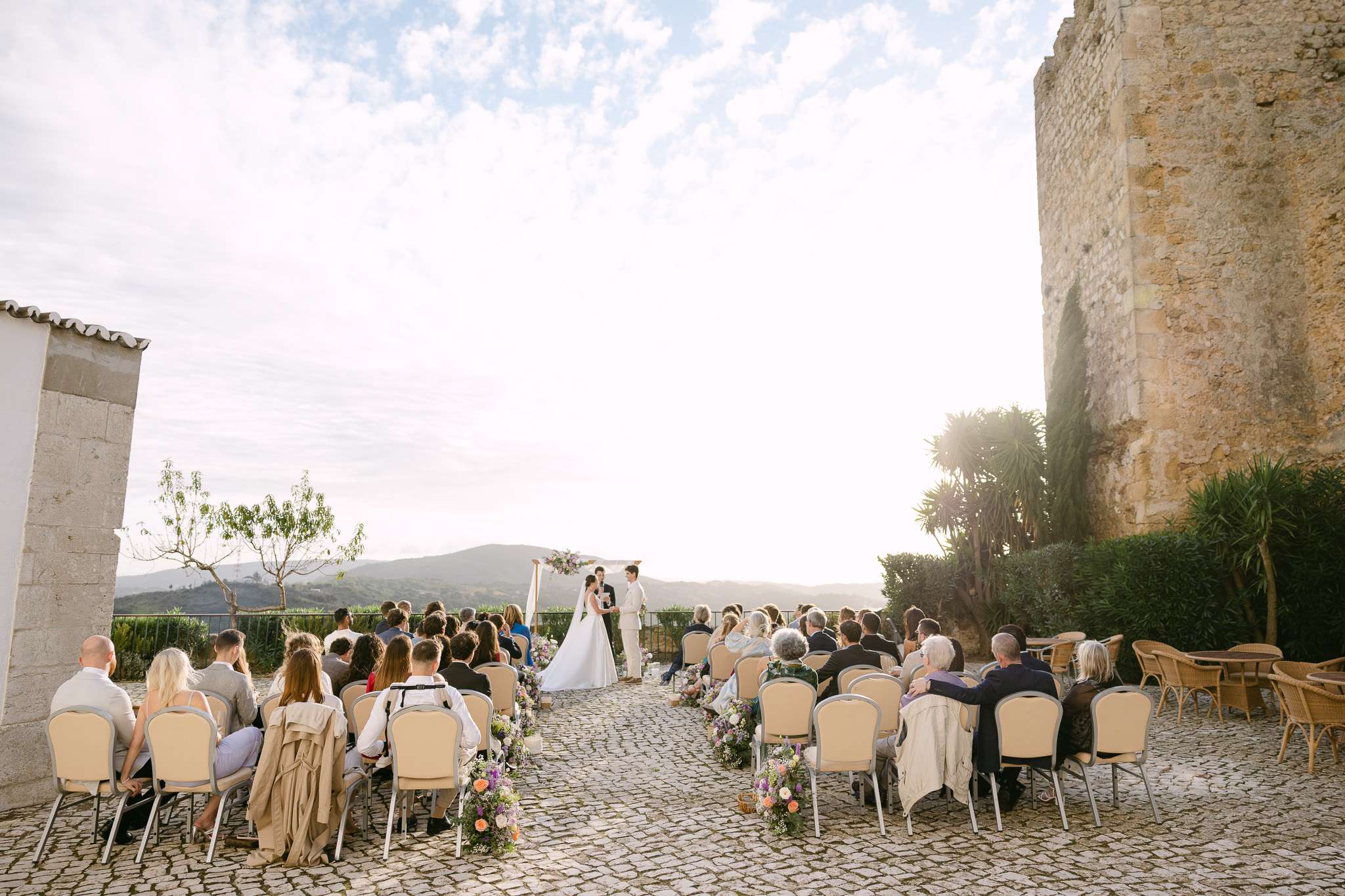 An outdoor pousada do castelo de palmela wedding ceremony with guests seated, facing a couple at the altar under a floral arch on a stone courtyard beside an old stone building, with scenic hills and a bright sky in the background.