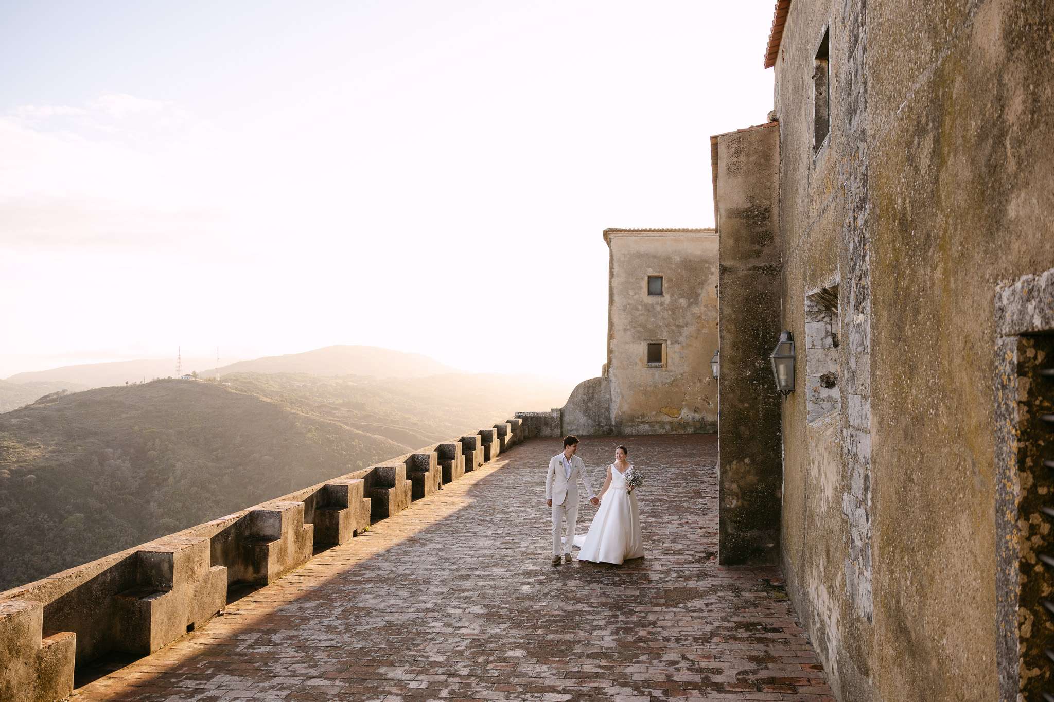 A bride and groom stand together on a large stone terrace beside an old building at Pousada do Castelo de Palmela, with mountains and a hazy sky in the background, bathed in warm, golden sunlight.