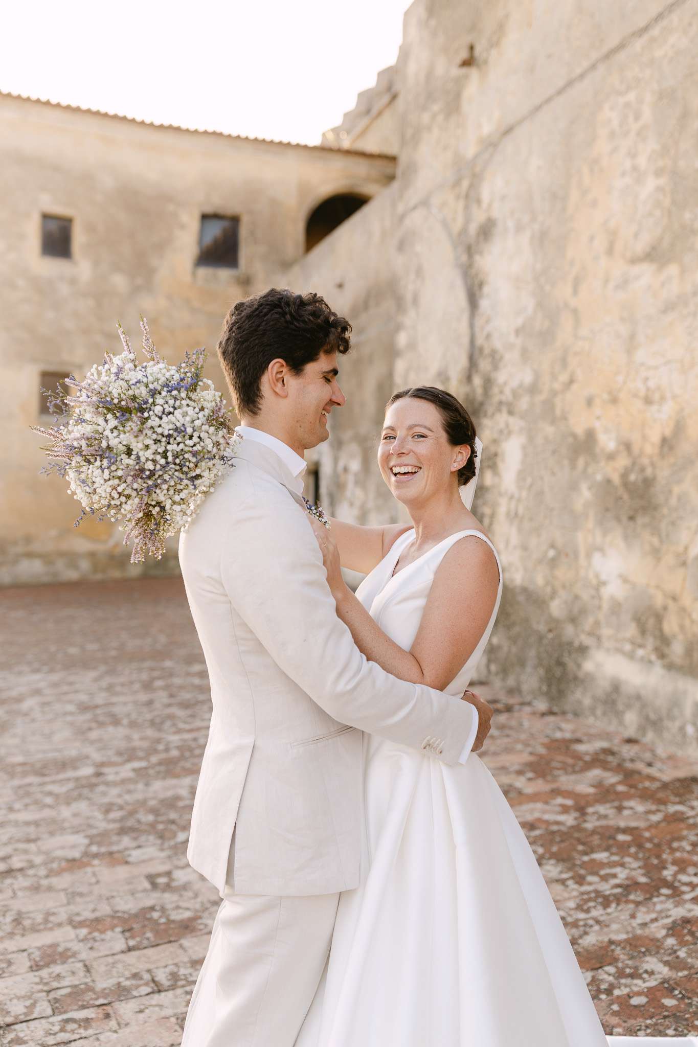 Uma noiva e um noivo em trajes brancos sorriem e se abraçam ao ar livre contra uma parede de pedra antiga e texturizada. O noivo segura um buquê de pequenas flores brancas sobre o ombro; o vestido da noiva é elegante e sem mangas.