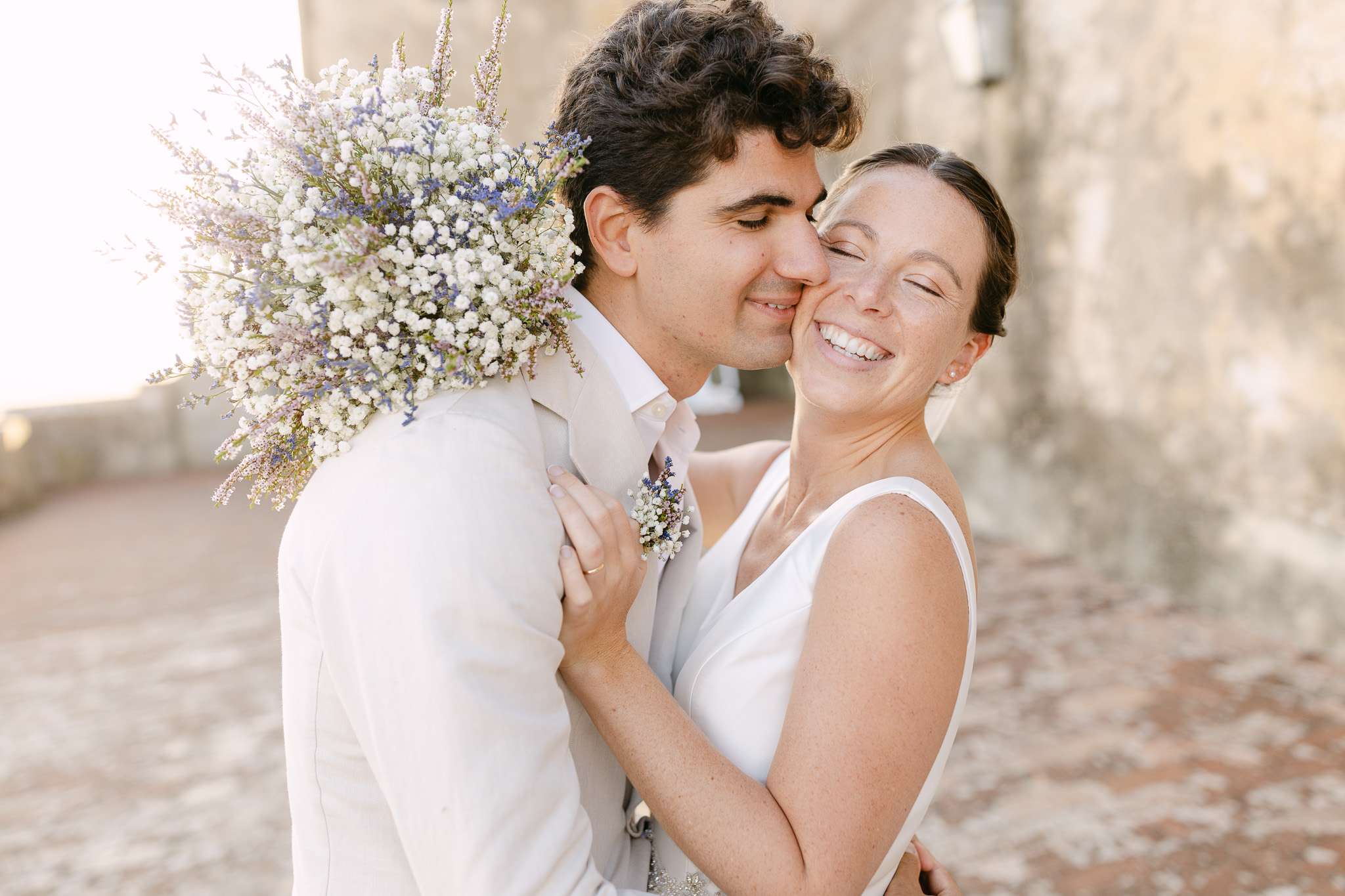 A bride and groom in white outfits embrace and smile joyfully outdoors at a Pousada do Castelo de Palmela wedding, with the groom holding a bouquet of white and purple flowers over his shoulder against a sunlit stone background.