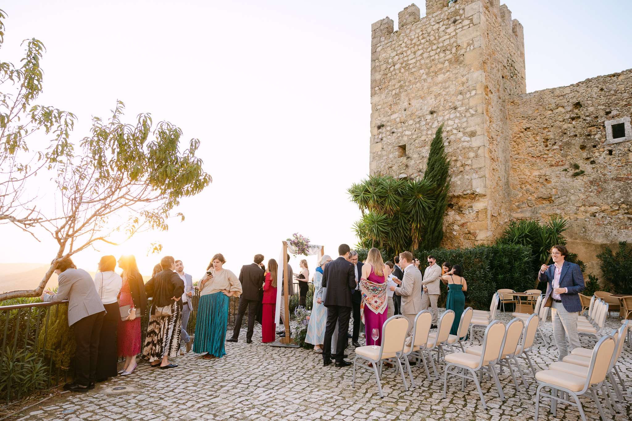 A group of people in formal attire gather outdoors near the Pousada do Castelo de Palmela at sunset, some standing by a railing with empty chairs arranged nearby on a cobblestone patio, creating an elegant wedding atmosphere.