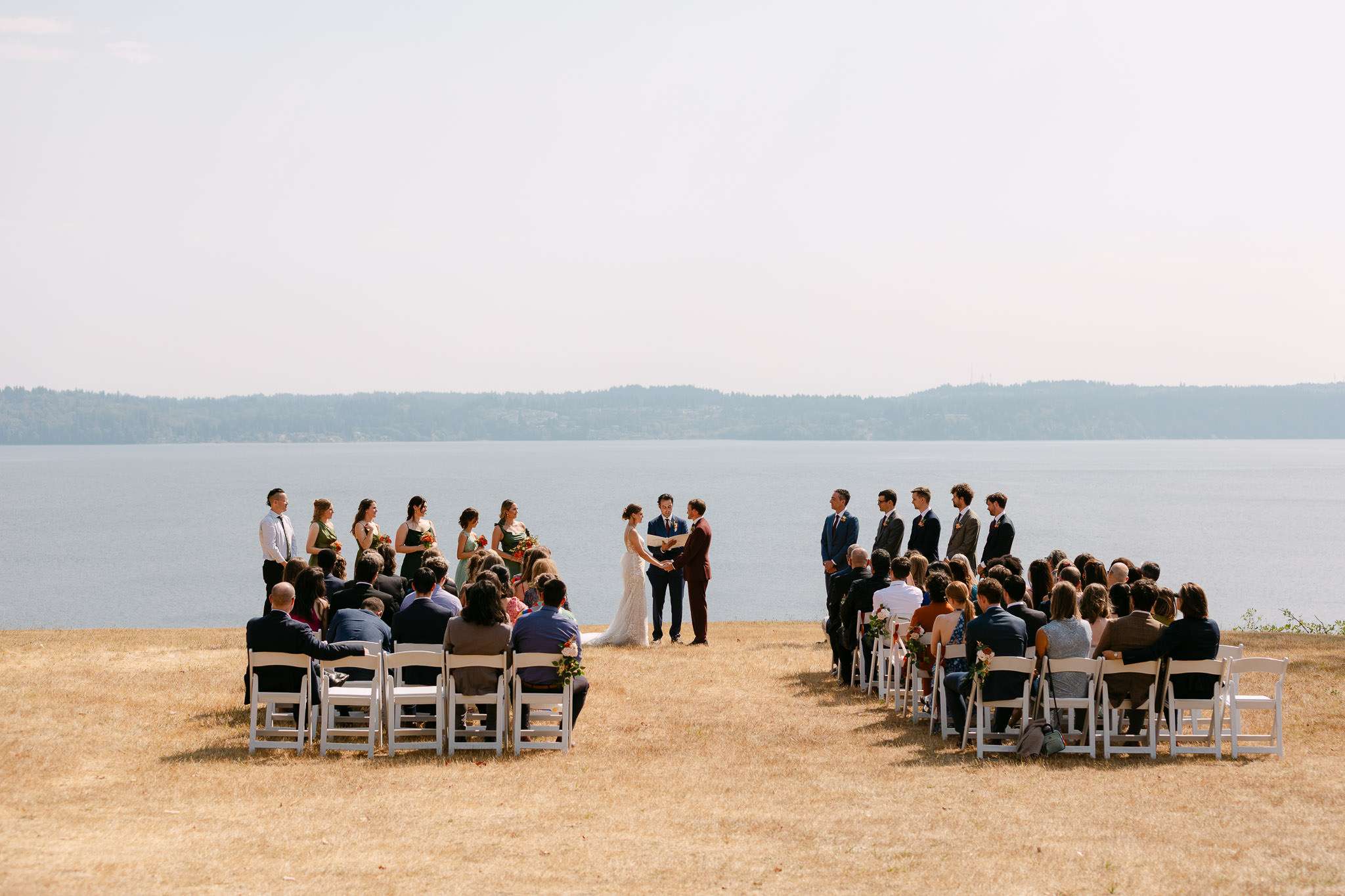 Outdoor wedding ceremony on golden clifftop overlooking a wide bay, bride and groom exchanging vows surrounded by wedding party and seated guests, destination wedding photography in portugal