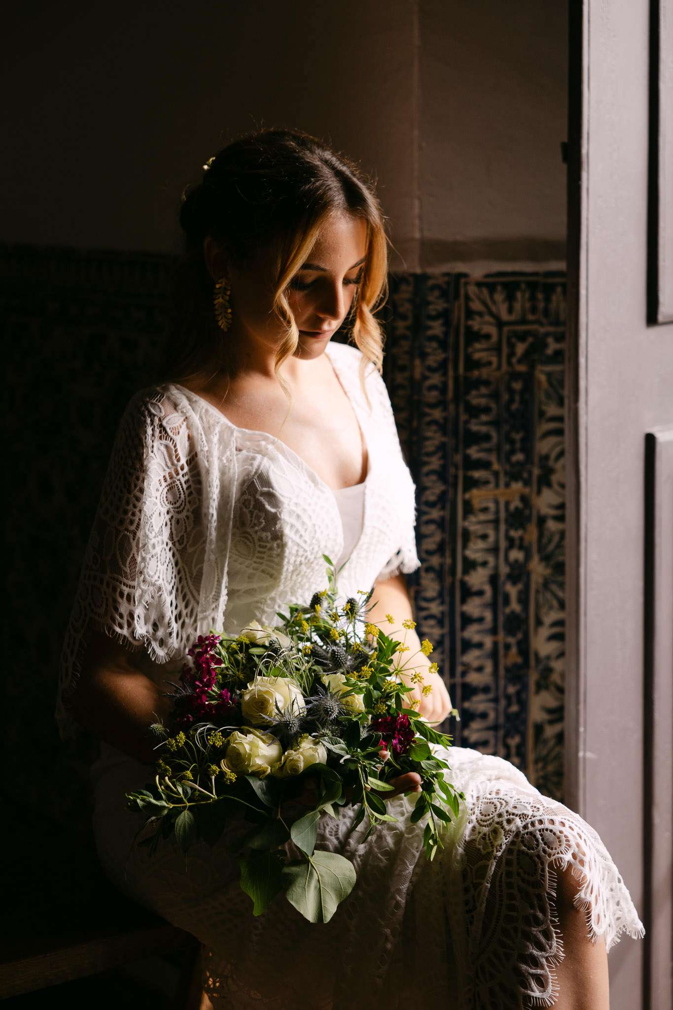 Bride in lace cape dress holding wildflower bouquet by window with azulejo tiles, documentary wedding photography Portugal