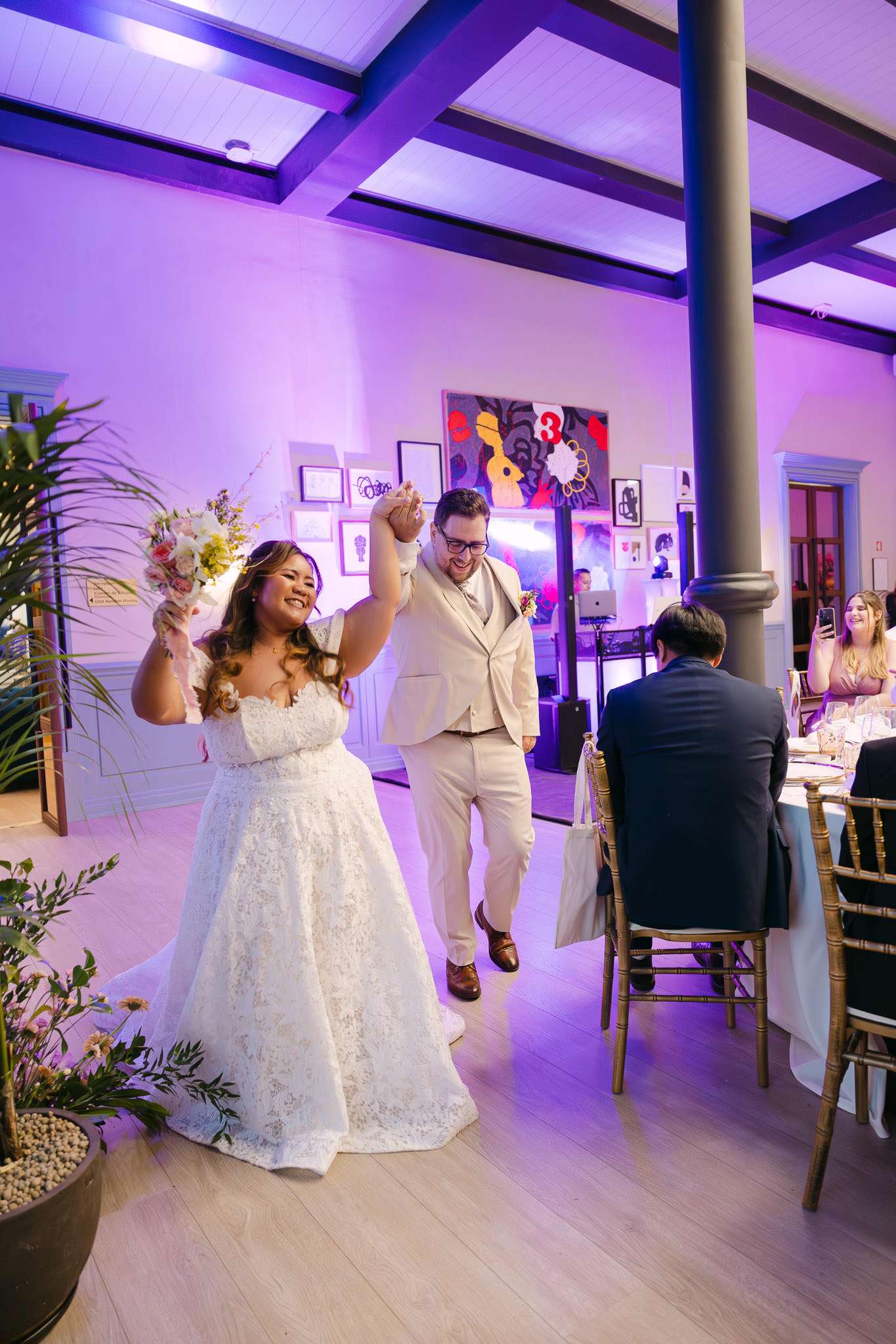 Bride and groom making joyful entrance into reception hall with purple lighting, bride raising bouquet, editorial documentary wedding photographer Portugal