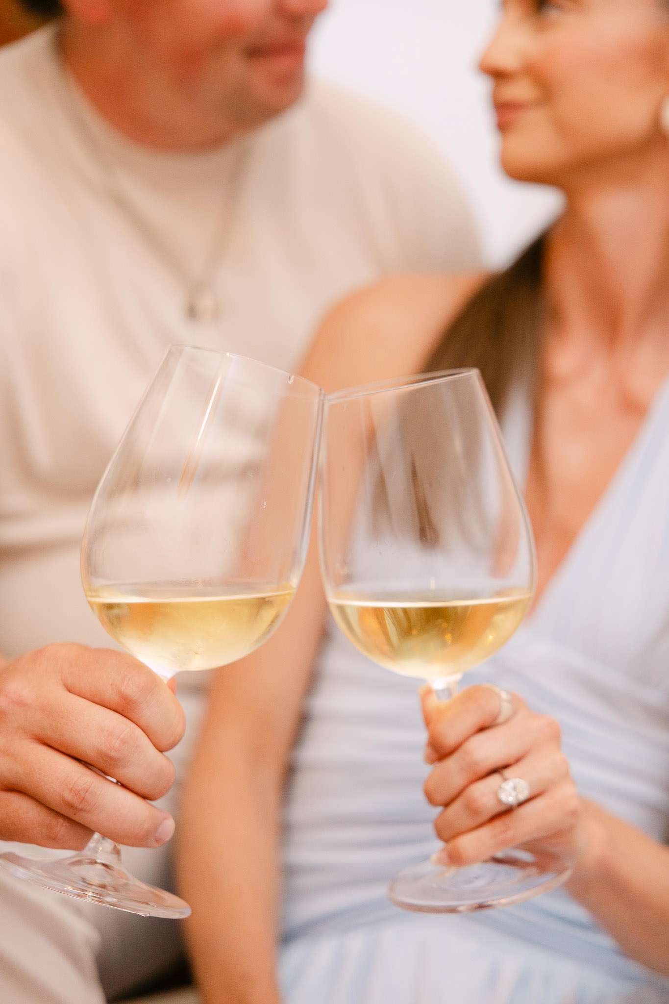 Two white wine glasses clinking at Comporta villa terrace, couple soft-focused behind, family holiday photography Portugal