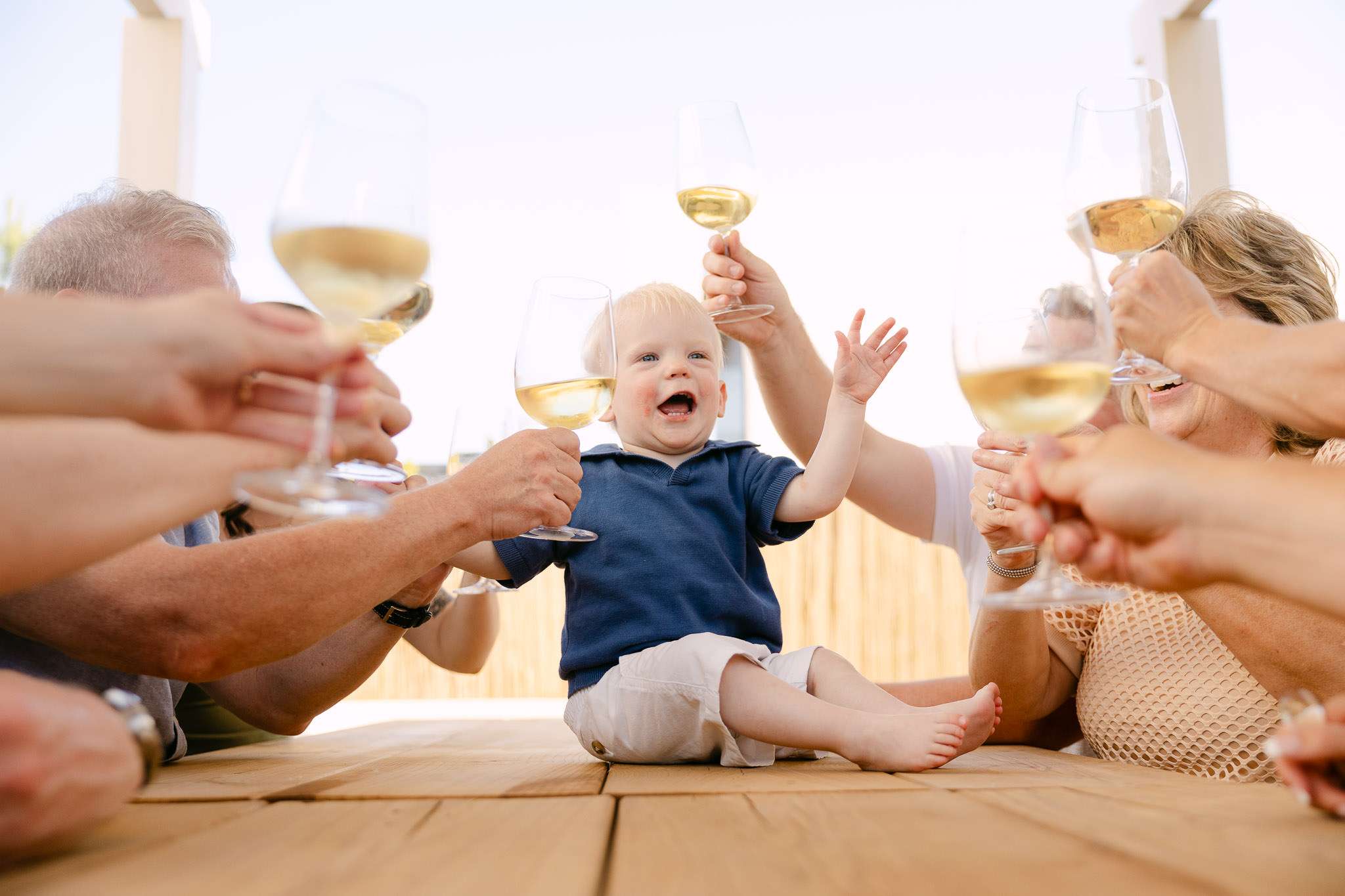 Baby reaching for wine glasses during family toast at Comporta villa terrace, laughing, documentary photography Portugal