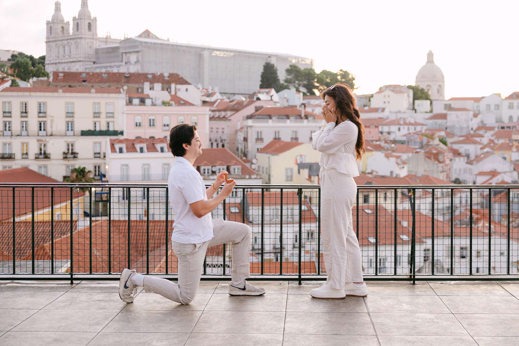Man on one knee proposing to a woman overcome with emotion at Portas do Sol viewpoint, with Alfama rooftops and São Vicente de Fora church in the background, Lisbon, Portugal