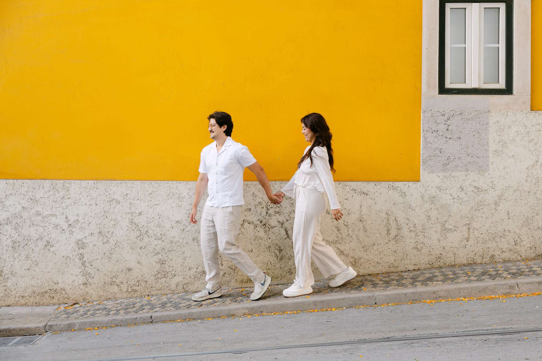 Engaged couple walking hand in hand past a bold yellow building wall on a cobblestone street in Alfama, Lisbon, candid proposal photography