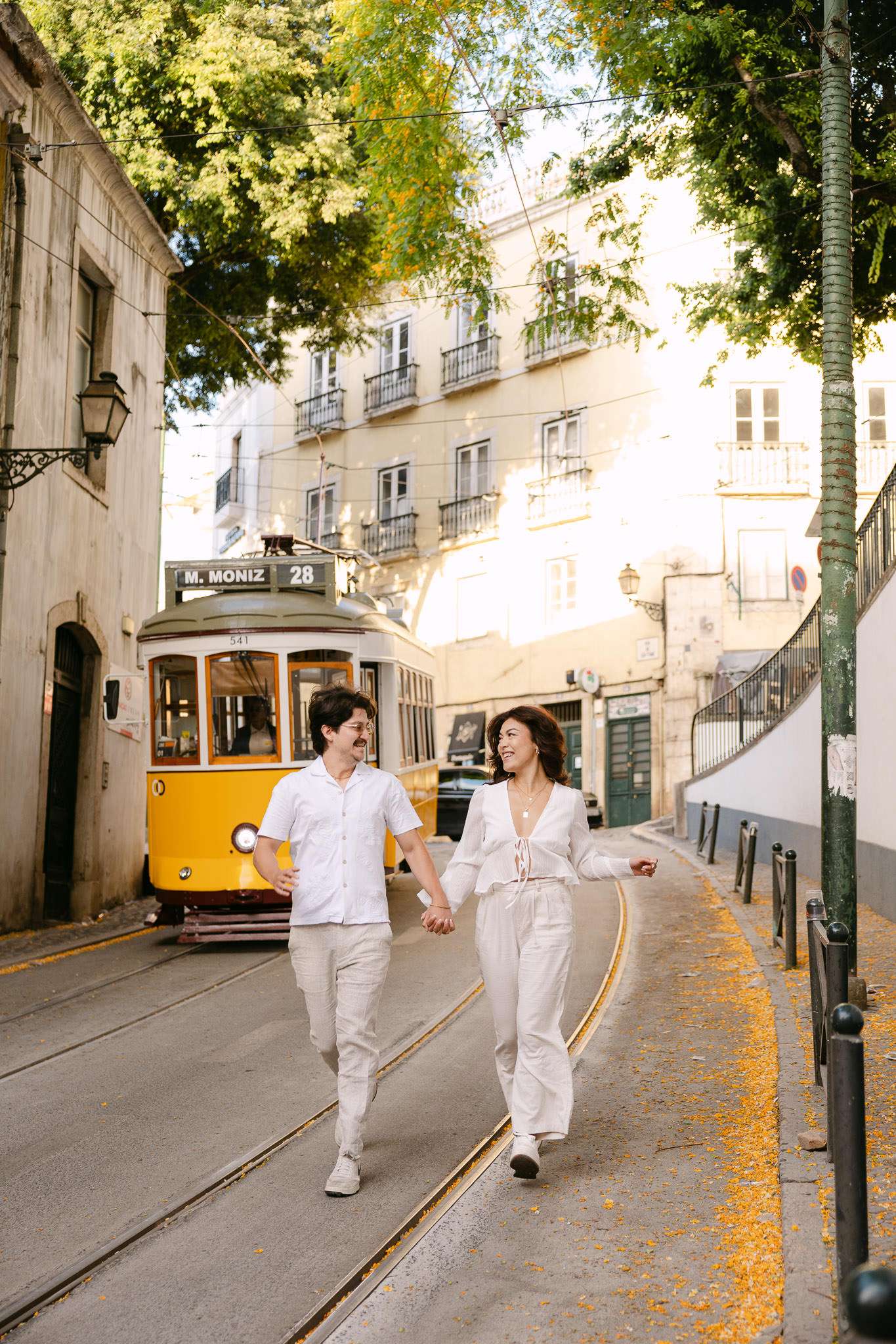 couple walking hand in hand and smiling at each other on a cobblestone street in Alfama with Lisbon's yellow tram 28 and golden morning light behind them, Lisbon proposal photography