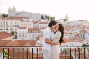 Newly engaged couple sharing their first kiss as a couple at Portas do Sol viewpoint, iconic Alfama rooftops and São Vicente de Fora church behind them, Lisbon, Portugal