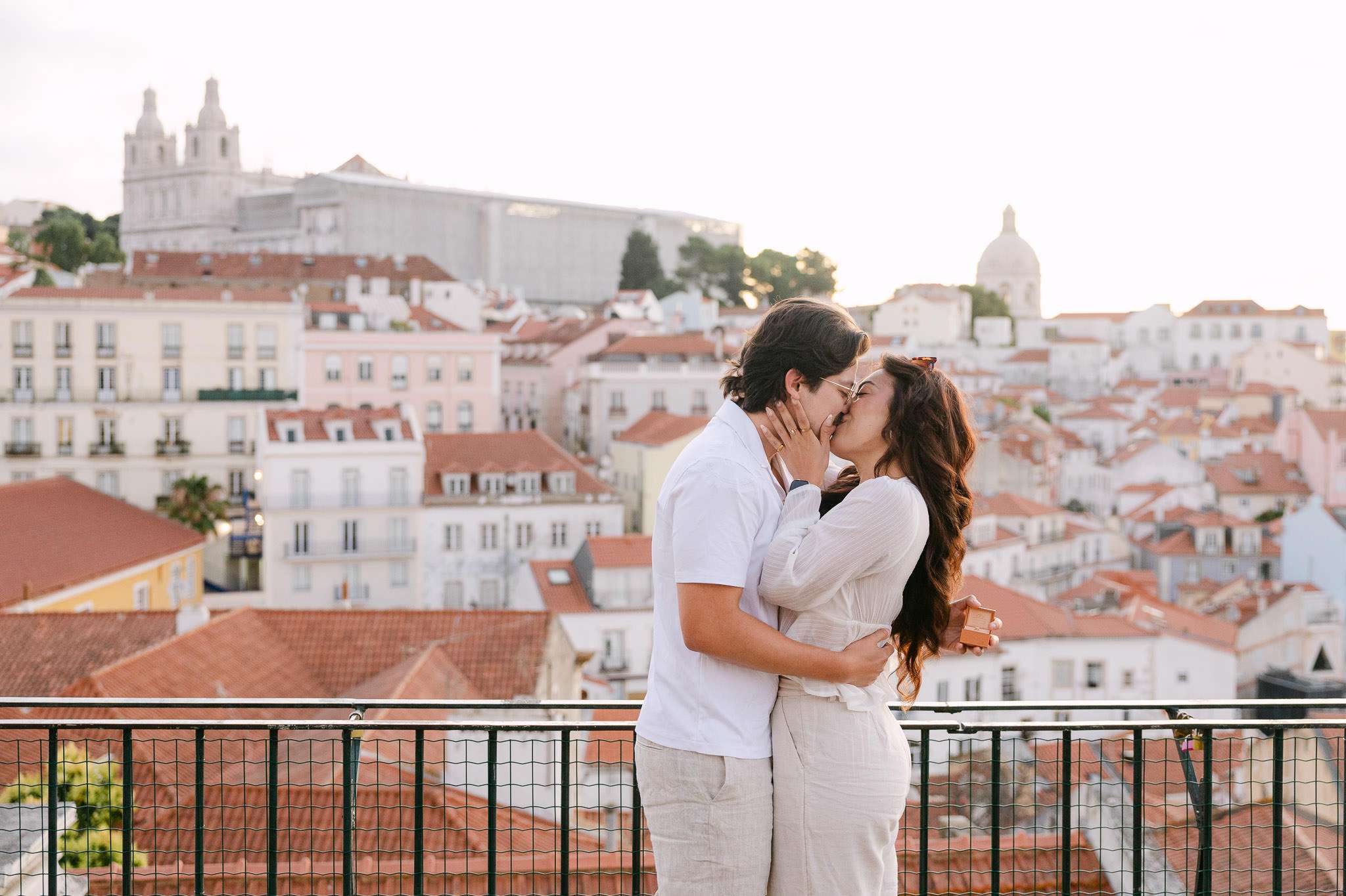 Newly engaged couple sharing their first kiss as a couple at Portas do Sol viewpoint, iconic Alfama rooftops and São Vicente de Fora church behind them, Lisbon, Portugal