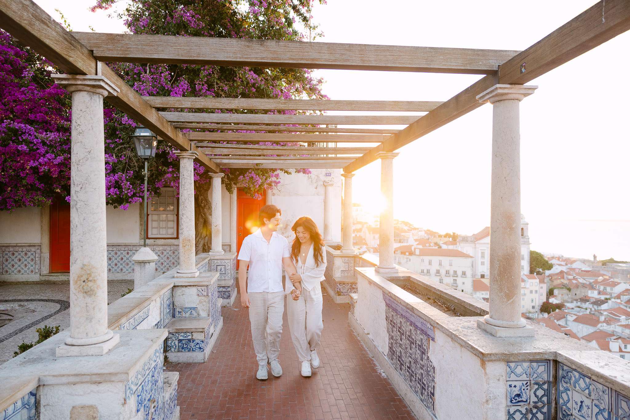 Engaged couple walking hand in hand under a pergola with vibrant purple bougainvillea and traditional azulejo tile benches at Miradouro de Santa Luzia, golden sunset light, Alfama, Lisbon