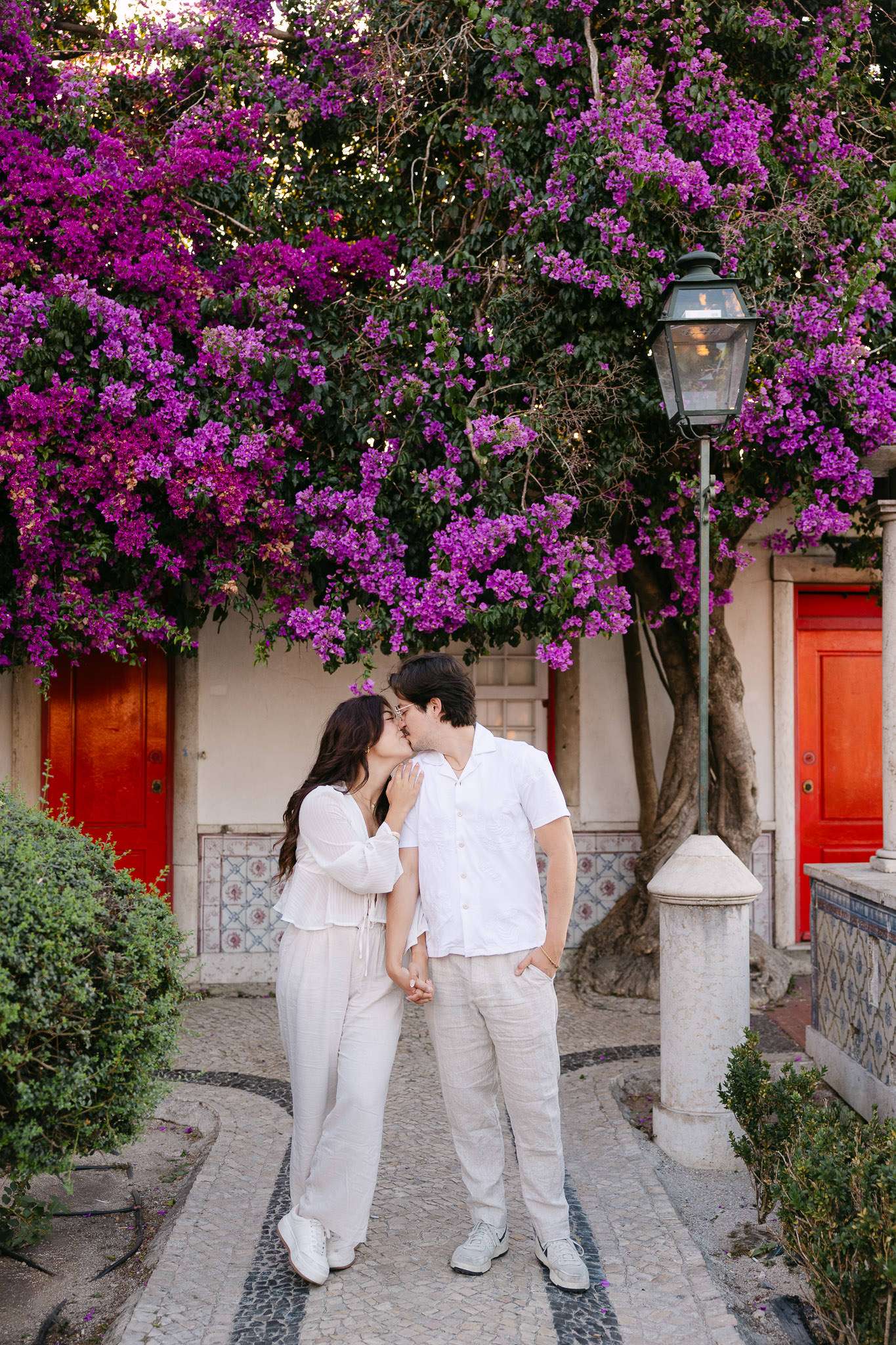 Couple kissing under an enormous purple bougainvillea tree with traditional red doors and azulejo tiles, cobblestone mosaic path, Miradouro de Santa Luzia, Alfama, Lisbon