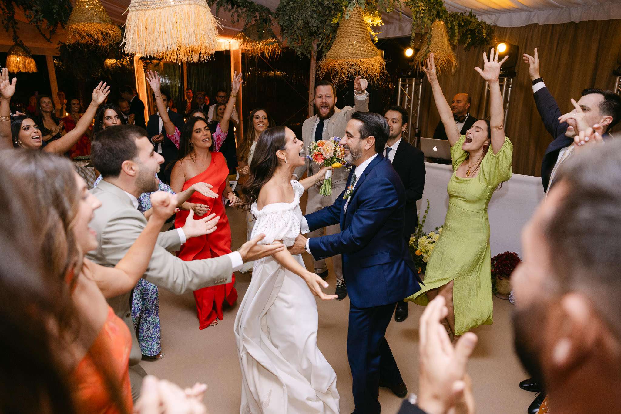 Bride and groom dancing surrounded by cheering guests with arms raised, joyful wedding reception under raffia pendant lights