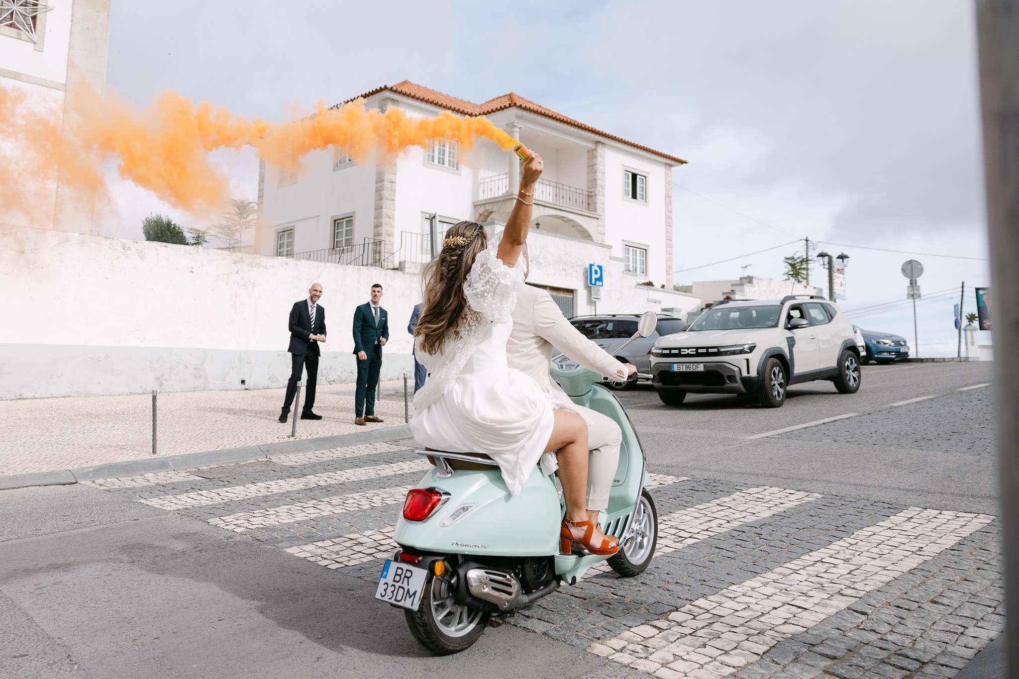 Bride holding orange smoke flare on Vespa scooter with groom through Palmela village streets, documentary wedding photographer Portugal