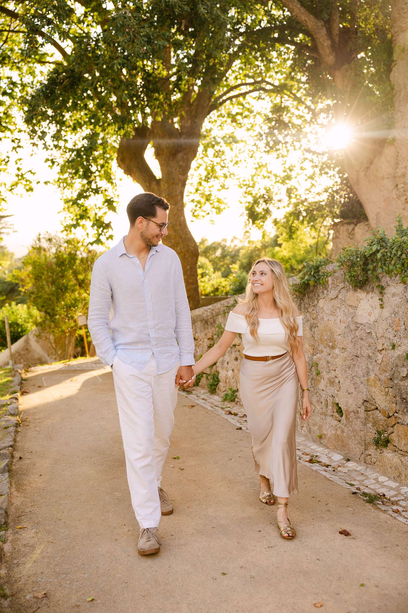 Engaged couple walking hand in hand along a garden path beside a stone wall, golden sunlight streaming through tree canopy, Sintra, Portugal proposal photography
