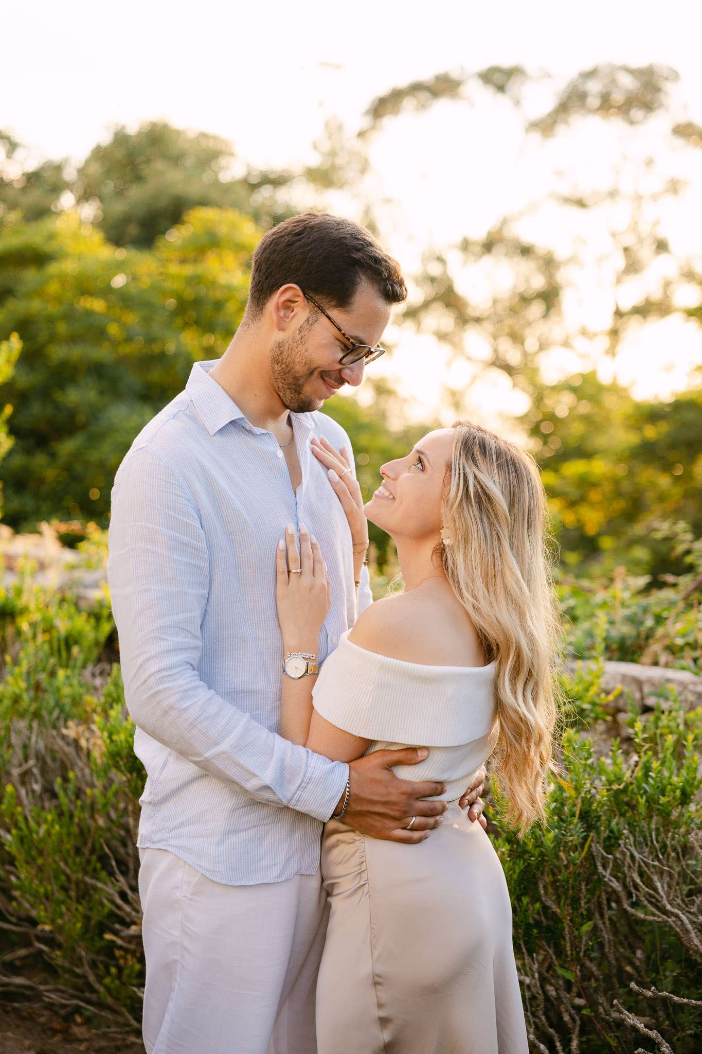 Newly engaged couple gazing at each other in warm golden hour light, woman's hand with engagement ring resting on man's chest, lush garden background, Sintra, Portugal