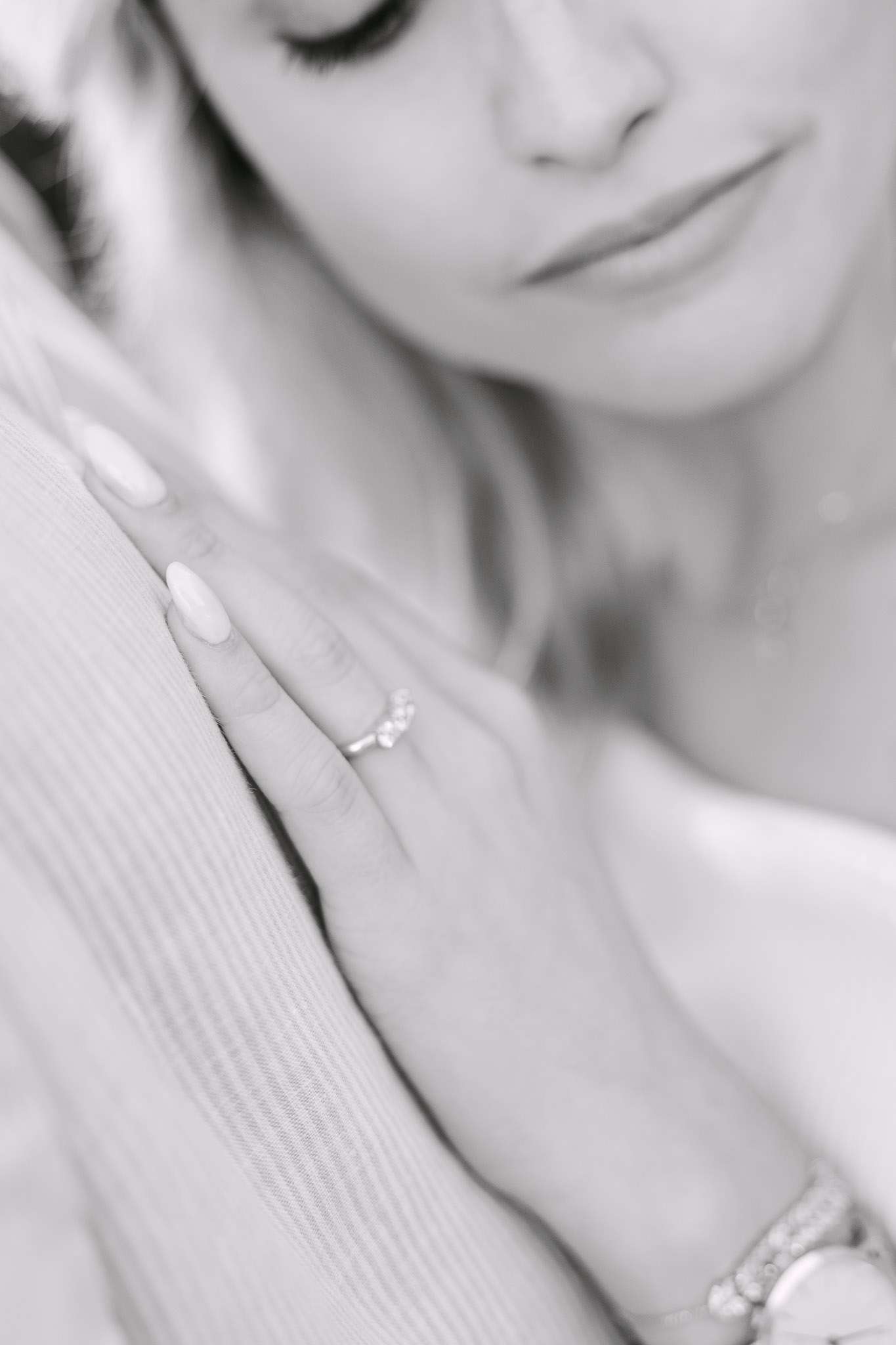 Black and white close-up of woman's hand showing diamond engagement ring resting on man's chest, her face softly blurred in background, intimate proposal photography detail