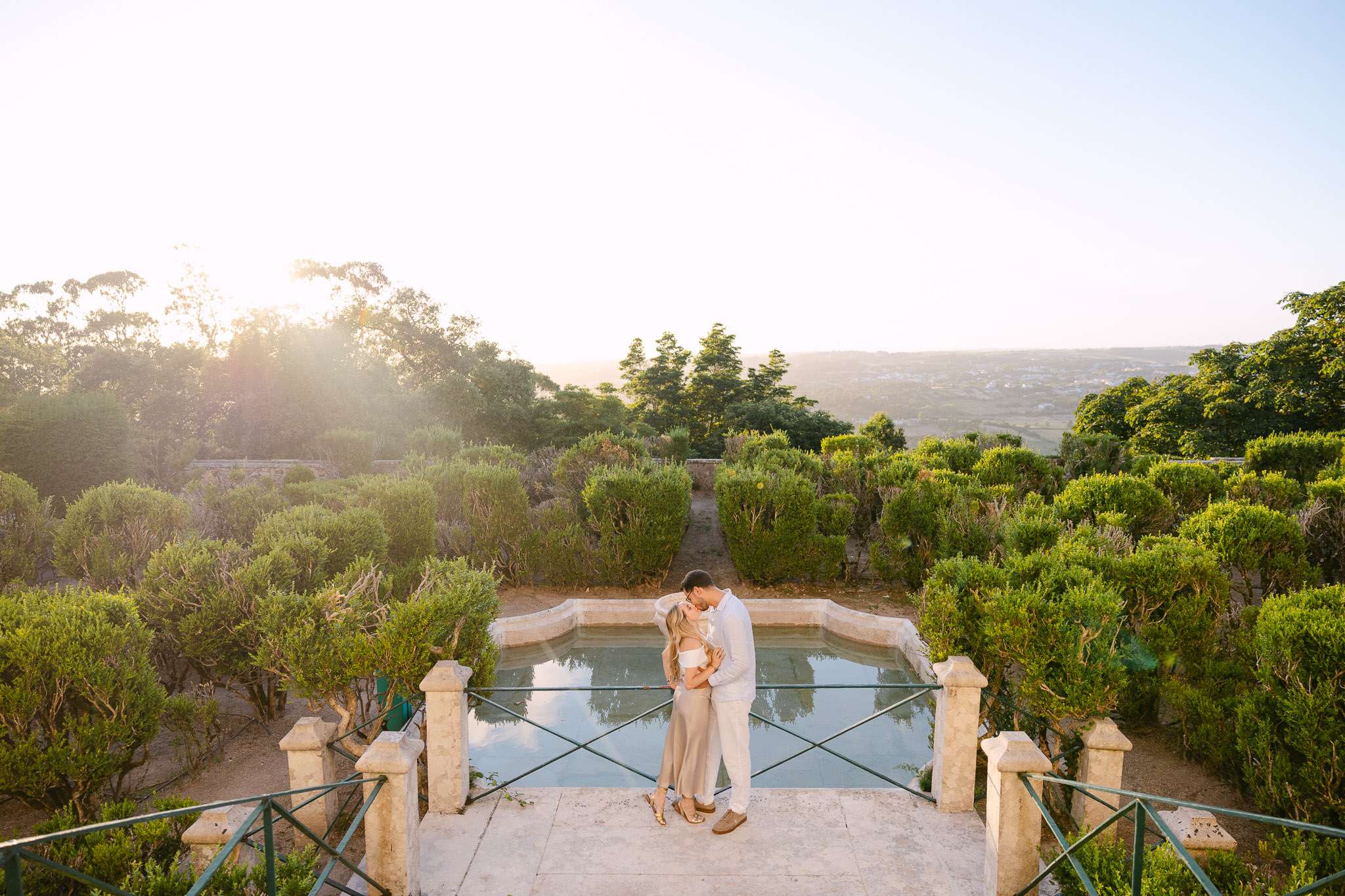 Couple kissing in front of an ornamental reflecting pool surrounded by manicured hedges, with a sweeping panoramic view of the Portuguese countryside at golden hour, Sintra