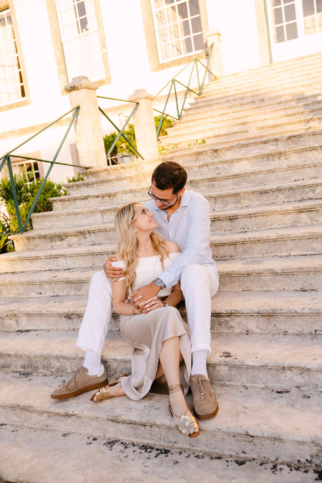 Engaged couple sitting together on wide stone palace steps, woman looking up at man smiling, warm golden light, Sintra, Portugal proposal photography