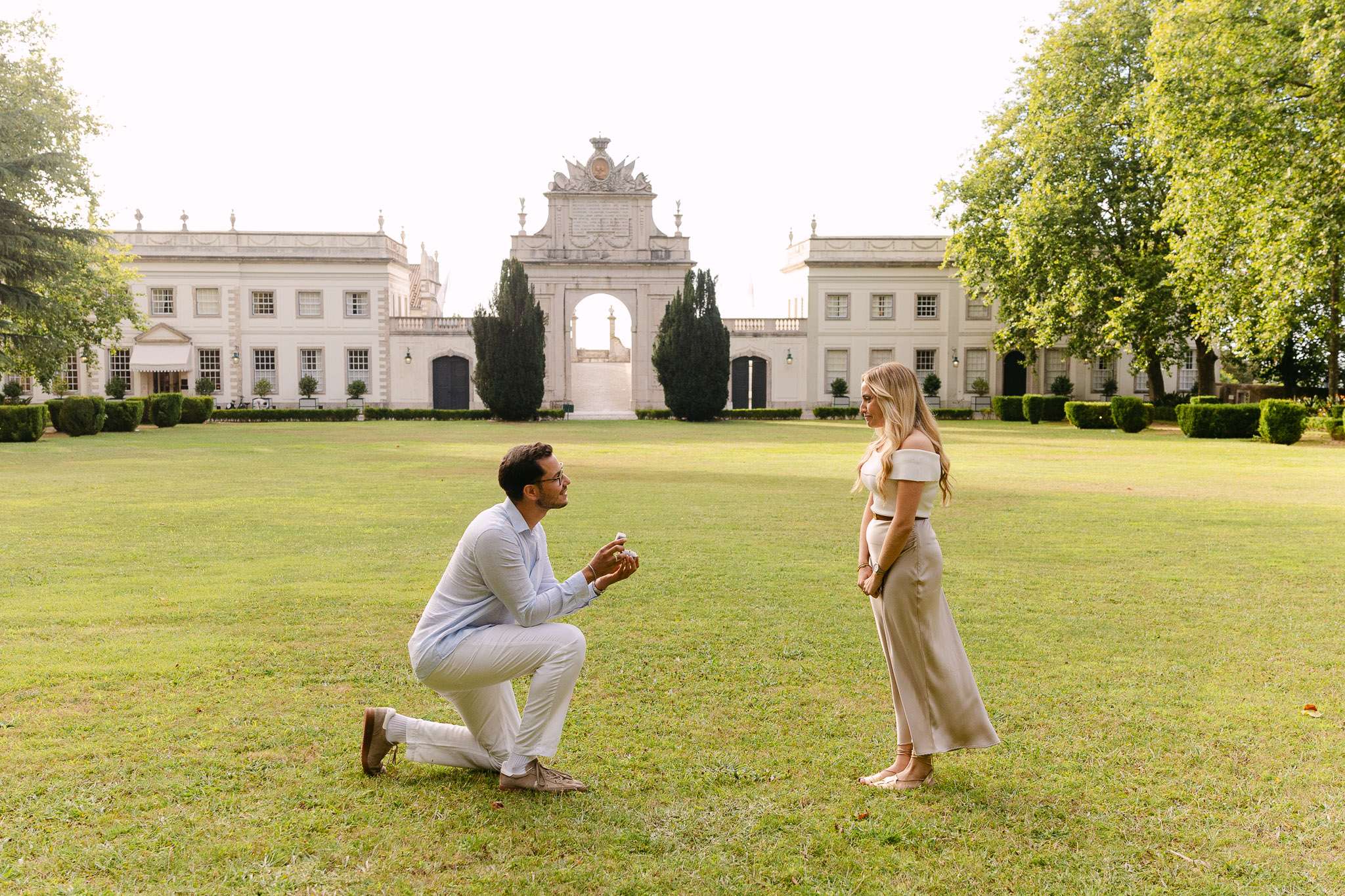 Man on one knee proposing to a woman on the green lawn in front of a white baroque palace in Sintra, Portugal, warm golden hour light