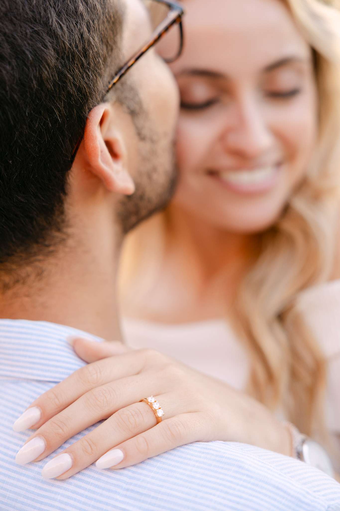 Close-up of woman's hand showing gold three-stone diamond engagement ring resting on man's shoulder, faces softly blurred in background, Sintra proposal photography