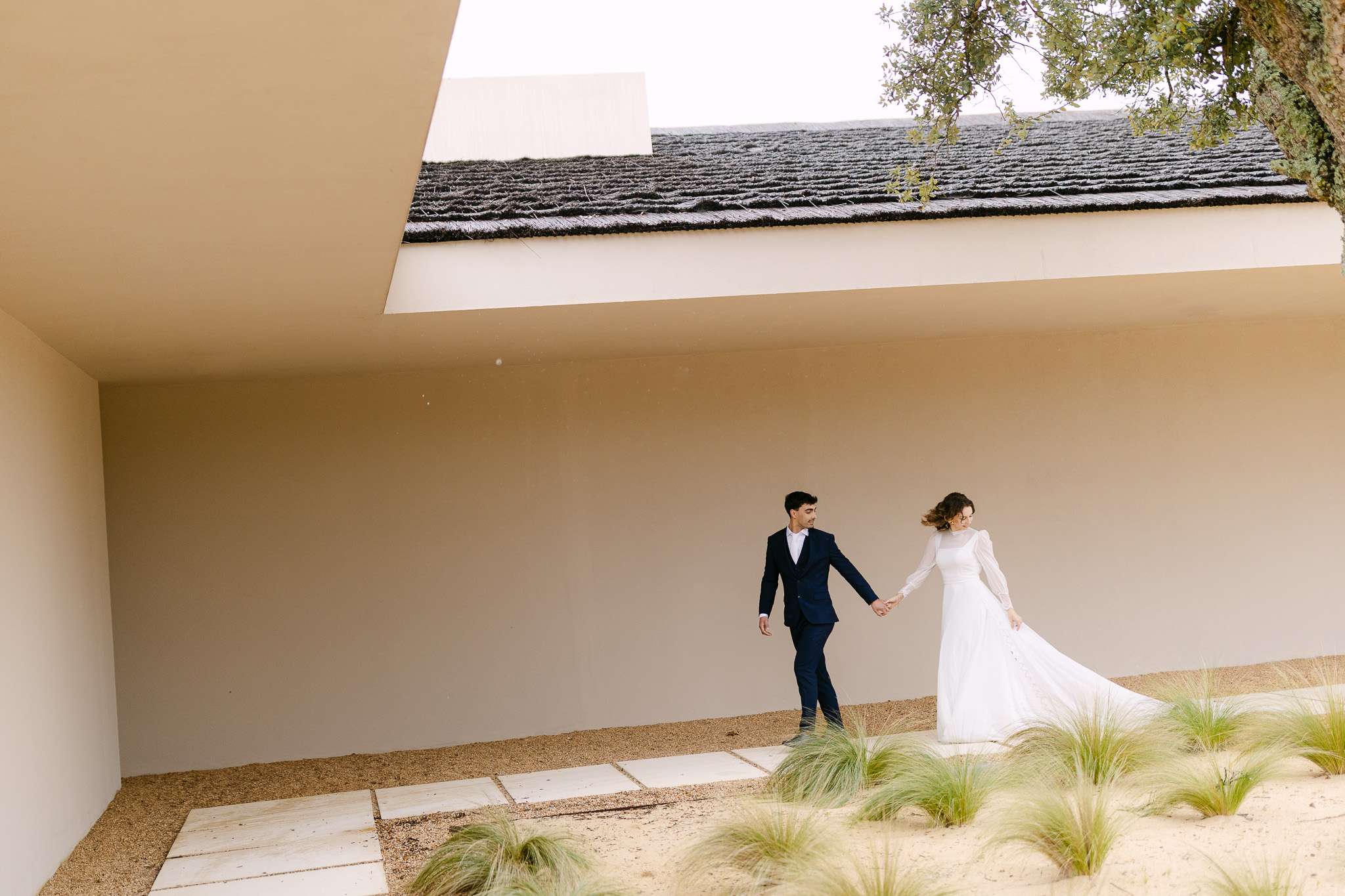 Comporta wedding photography of bride and groom walking hand in hand past a thatched-roof building.