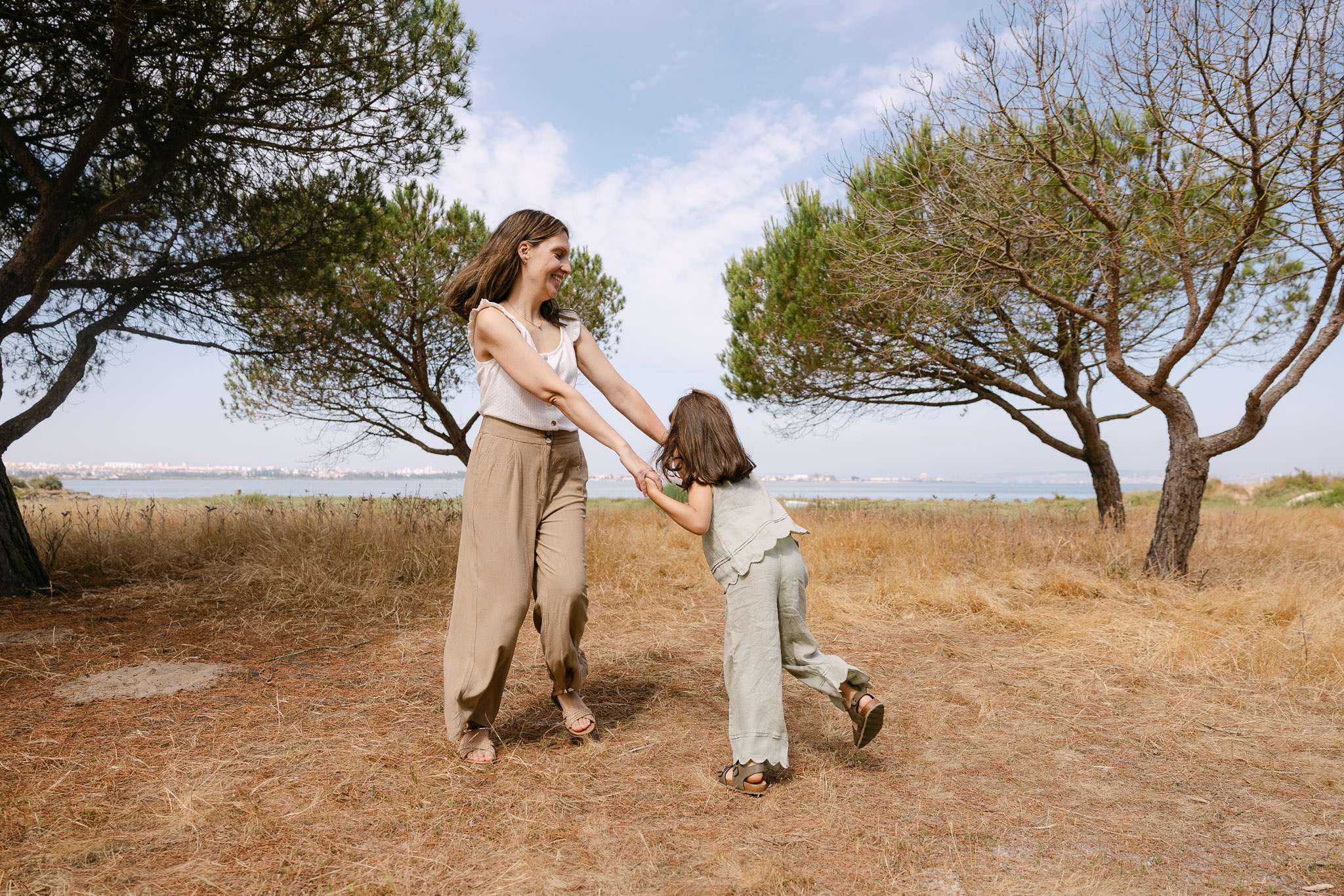 Mother and daughter spinning joyfully in a sun-dried meadow with pine trees and the Sado estuary in the background, family photoshoot Setúbal