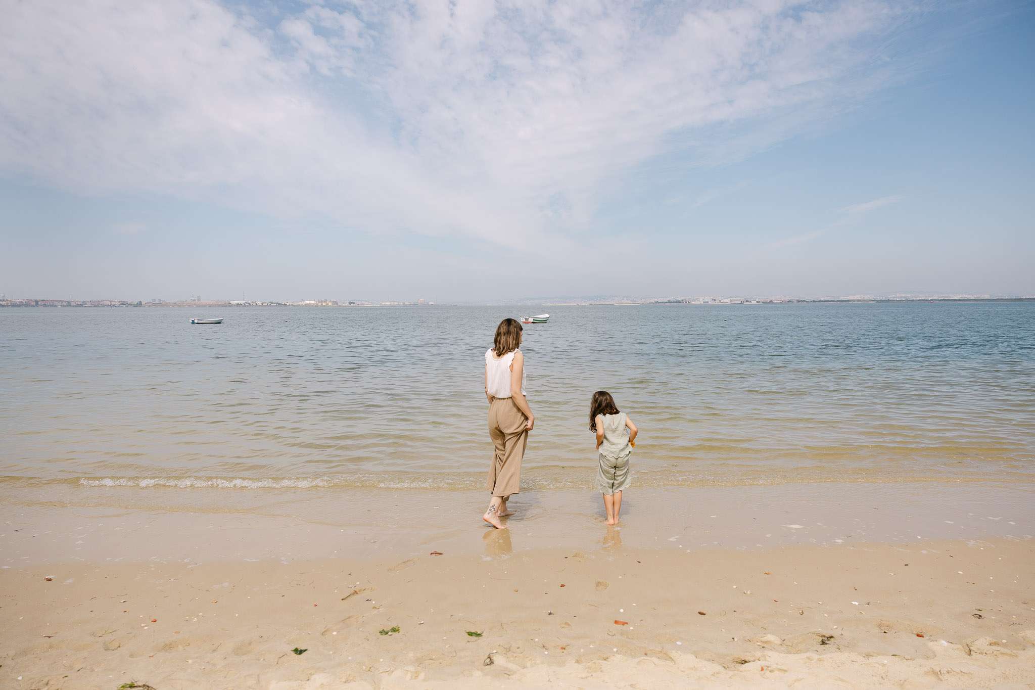 Mother and daughter standing barefoot at the water's edge on a calm beach, looking out at the estuary, family photography Setúbal Palmela